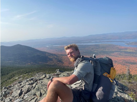 The presenter Brandon Weis is wearing a backpack and is seated on a rock with green and red valley behind him