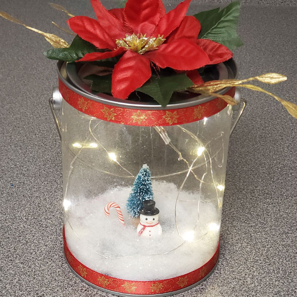A clear paint can with a pine tree, snowman, and candy cane inside lit up by fairy lights. On top of the paint can is a poinsettia arrangement.