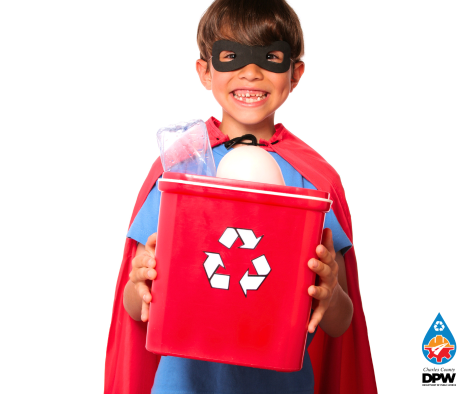 Smiling masked child in a red superhero cape holding a red recycling bin filled with items.