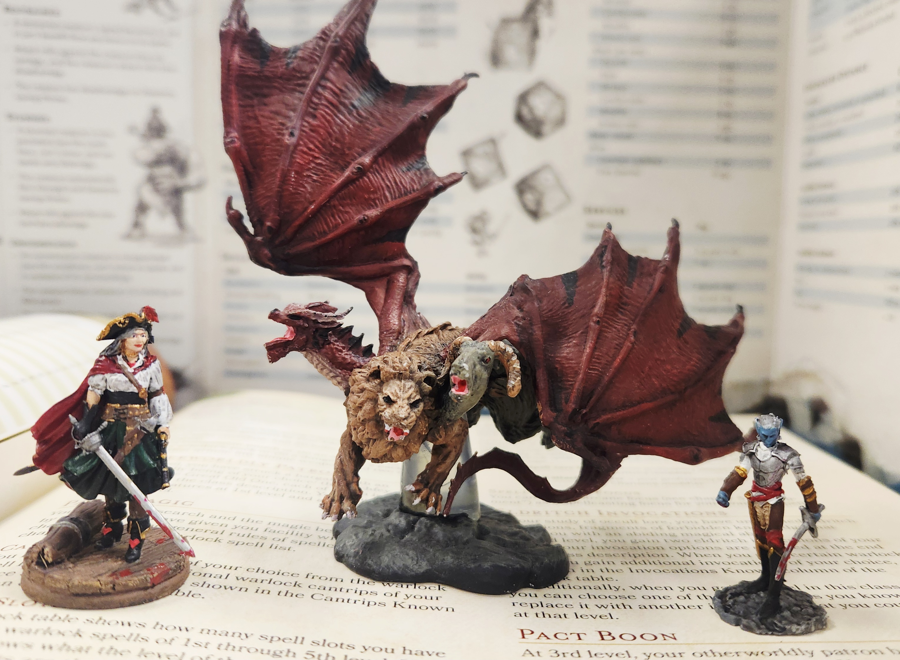 3 D&D Miniatures stand on top of a D&D rulebook. Left: a female pirate captain holding a bloody sword, Middle: a large chimera with the heads of a dragon, lion, and goat, Right: a pirate fighter holding a bloody sword.