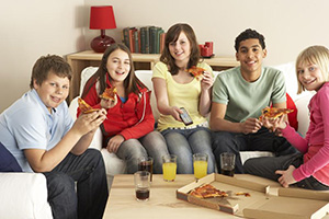 
Un grupo de cinco adolescentes sonrientes sentados en sofás y comiendo pizza