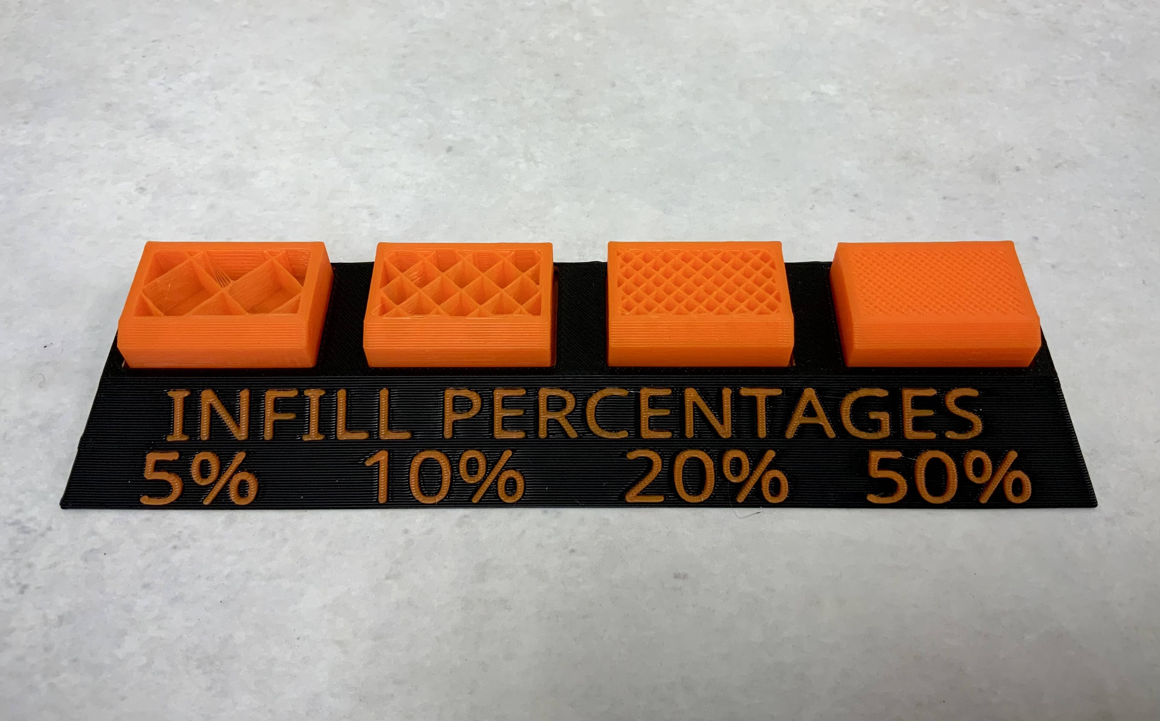 Four orange 3D printed blocks with exposed insides are side by side on a black plinth with orange text which reads "Infill Percentages" and the figures 5%, 10%, 20%, and 50% under each block. Each block has walls in a diagonal grid formation inside with different spacing. The left block has large squares of space, with each subsequent block showing progressively less negative space, until the right block whose squares are barely visible.