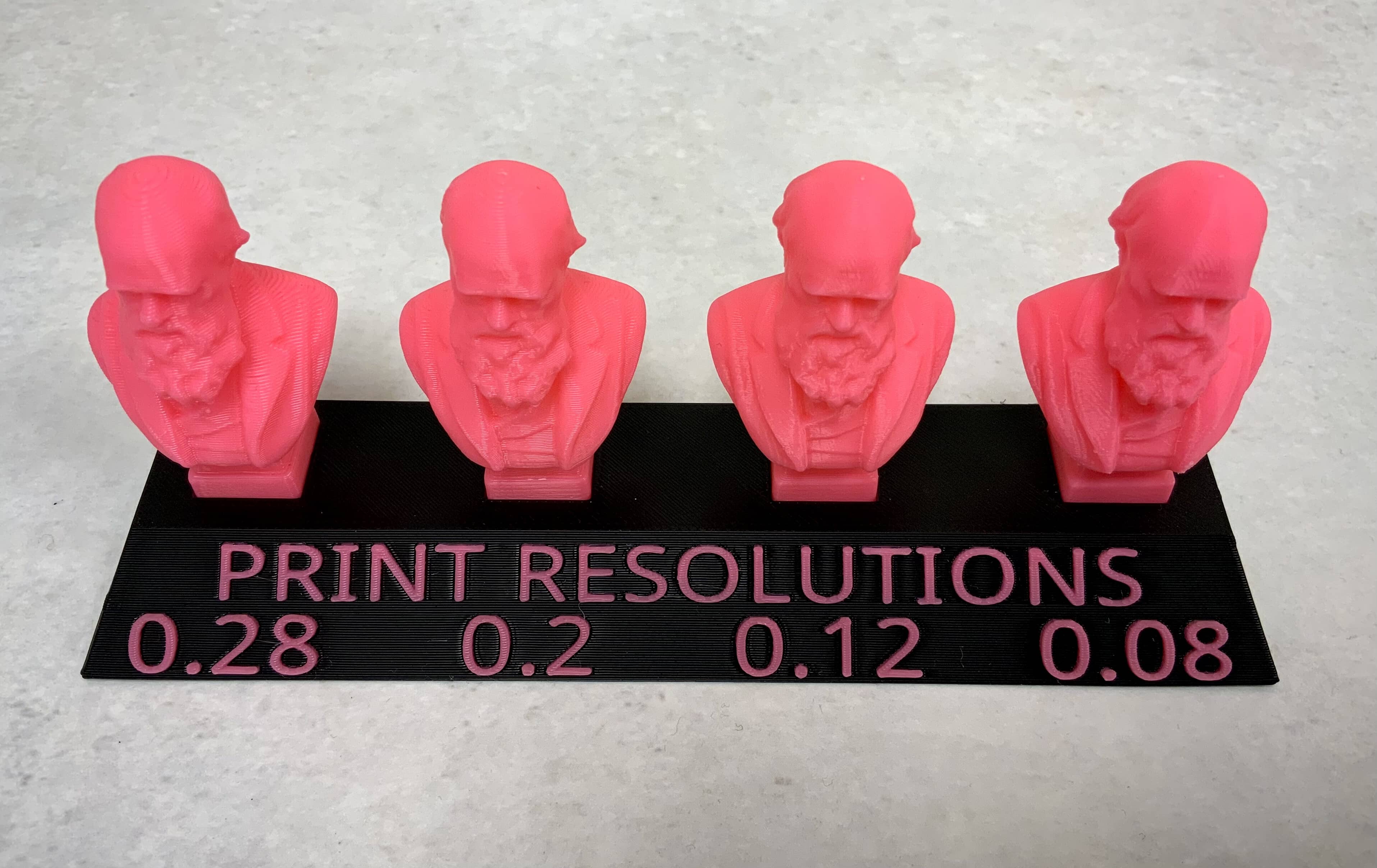 Four pink 3D printed busts of Charles Darwin side by side on a black plinth with pink text which reads "Print Resolutions" and the numbers 0.28, 0.2, 0.12, and 0.08 under each bust. The left bust has visible stair-stepped layers, with each subsequent bust showing subtler layers until the right bust which has barely visible layer lines.