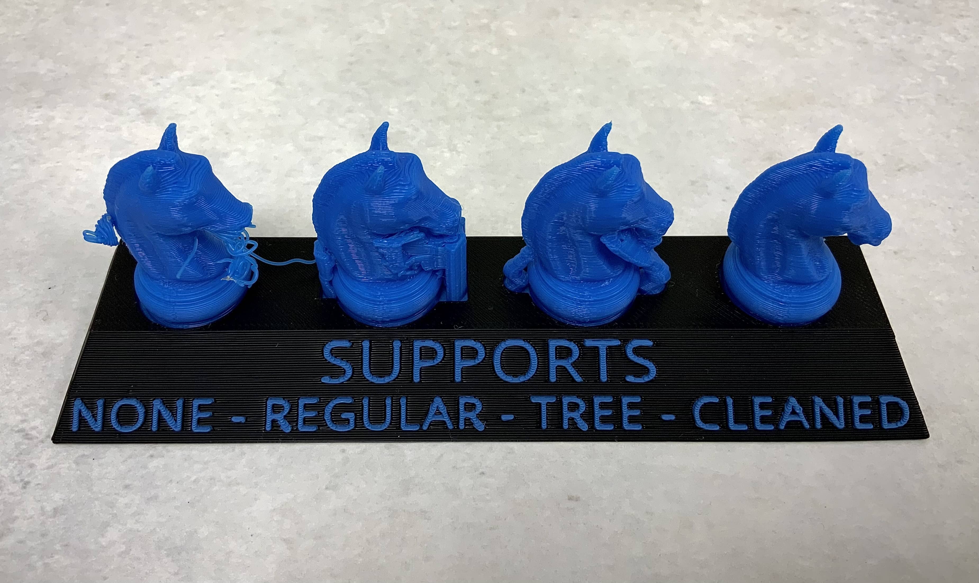 Four blue 3D printed chess set knights side by side on a black plinth with blue text which reads "Supports" and the words unsupported, regular, tree, and cleaned under each knight. The left knight has a snout akin to unraveled yarn, the regular knight has blue blocks under its snout and mane, the tree knight has thick blue branches supporting the snout and mane, while the cleaned knight shows a completed chess piece without issues.