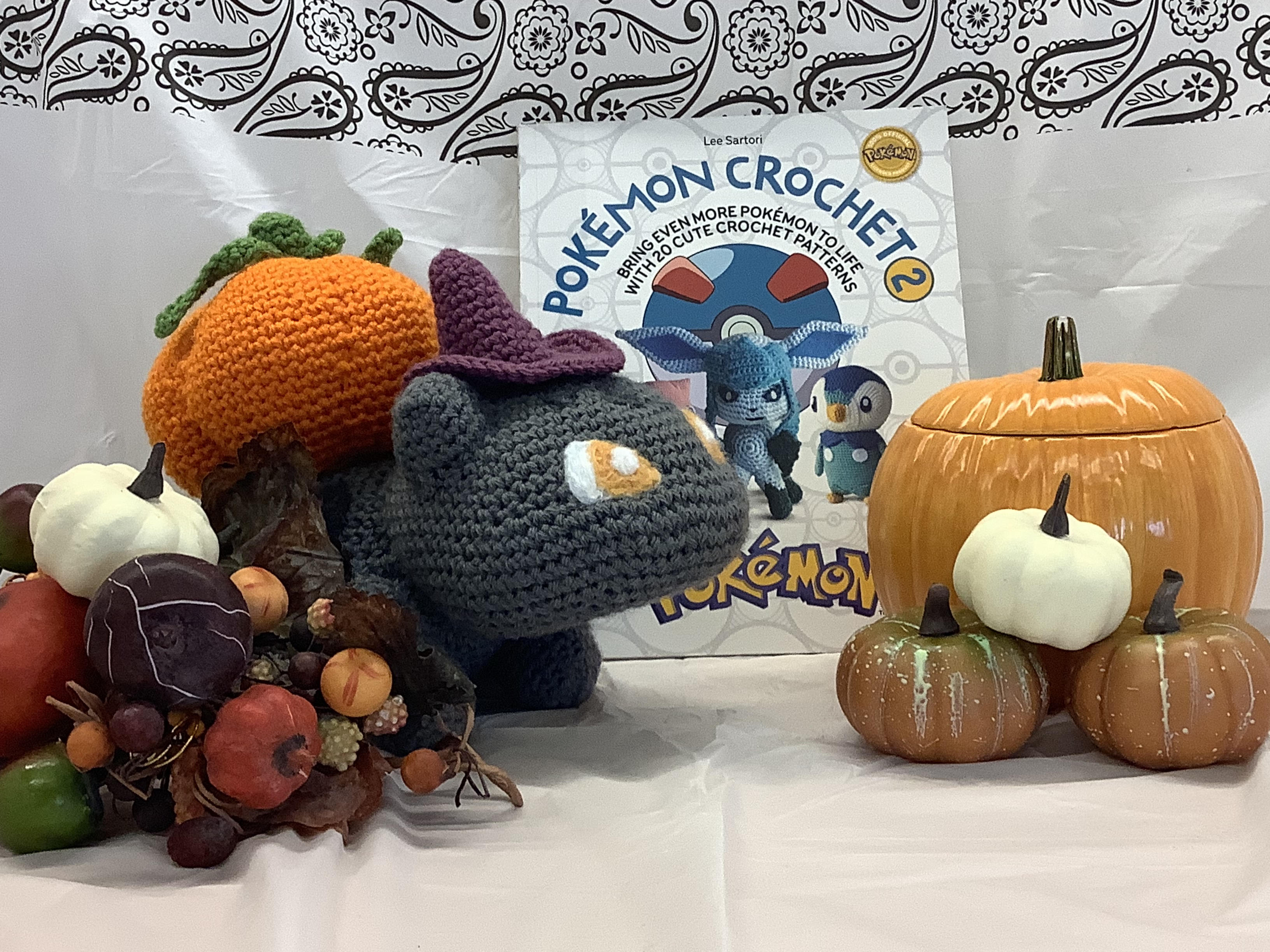 A large crocheted bulbasaur with a witch hat and some ceramic pumpkins next to a book titled Pokemon Crochet