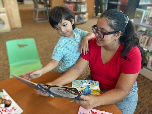 A mom with glasses and a long ponytail reads from a board book to a kindergartner wearing a striped shirt.