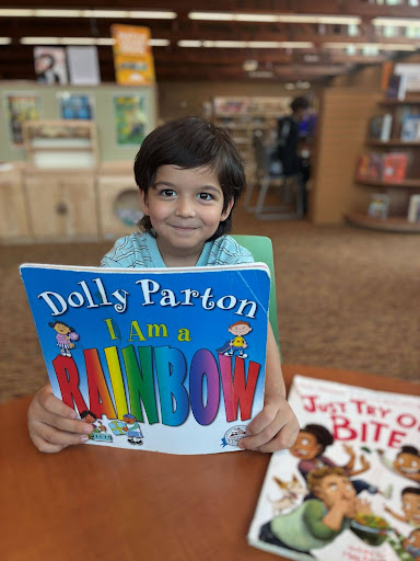A kindergartner in a striped shirt holds up Dolly Parton's I AM A RAINBOW while sitting at a Library table and smiling.