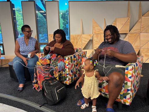 Three adults sit in colorful chairs crocheting with a toddler at their feet