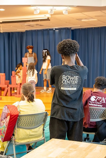 A teen in a Bowie Center T-shirt takes a photo of other teens on stage performing to preschoolers.