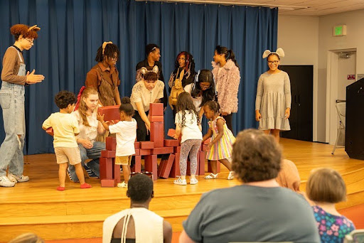 7 teens in animal headbands and 4 preschoolers build a block tower together on stage in front of a blue curtain while parents and caregivers watch from the audience