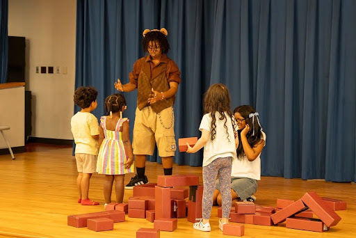A teen boy wearing bear ears engages with three Library preschoolers and a tumble of blocks on a stage.