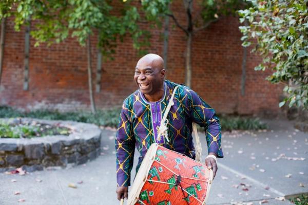 Onye wears a large drum over one arm and smiles as he stands outside in front of a wall and some small trees.