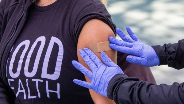 Gloved hands place a bandage on a person’s upper arm after an injection.